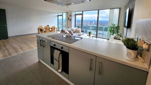 A modern kitchen with a large island, a dining area with a table and chairs, and a living space with a sofa and large windows offering a city view. - Stunning Penthouse with Balcony | Sunset Views - Pass the Keys (Belfast)