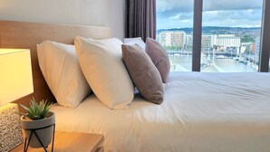 A hotel room with a bed, a bedside lamp, a small potted plant, and a view of the city through large windows. - Stunning Penthouse with Balcony | Sunset Views - Pass the Keys (Belfast)