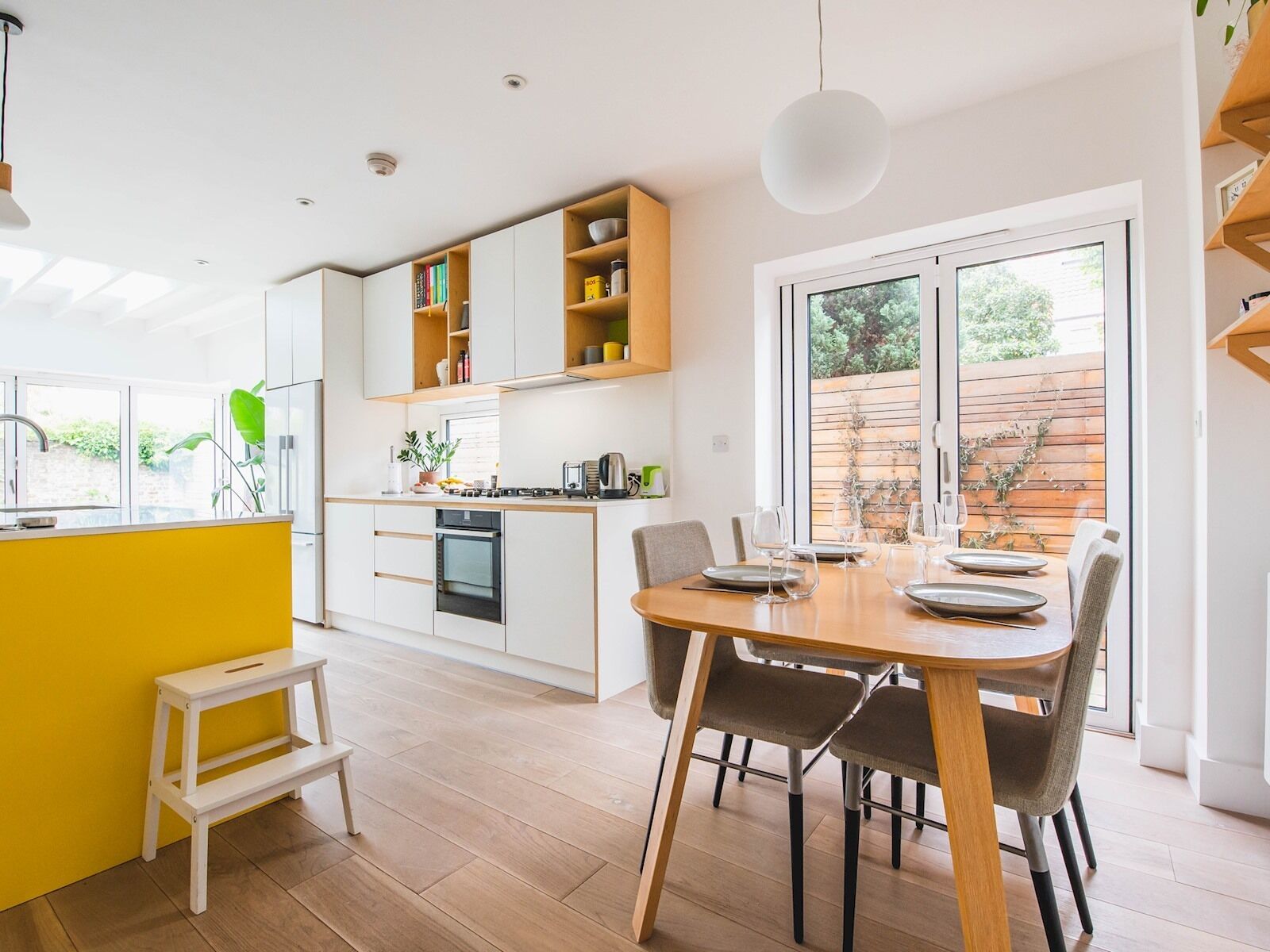 A modern kitchen with white cabinets, a yellow accent wall, and a dining area with a wooden table and chairs.