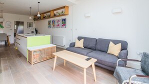Moderni olohuone, jossa on harmaa sohva, puinen sohvapöytä sekä keittiöalue, jossa on valkoinen työtaso ja puiset hyllyt.
