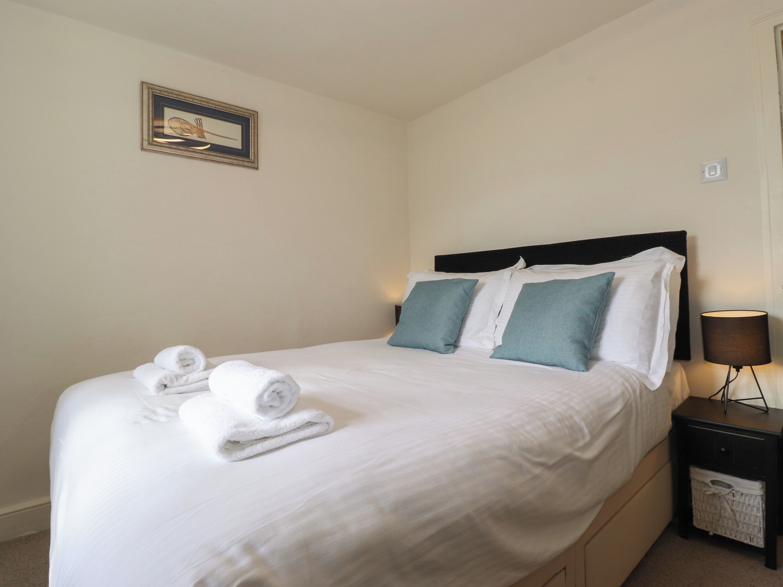 A neatly made bed with white linens and two pillows, a small bedside table with a lamp, and a framed picture on the wall.