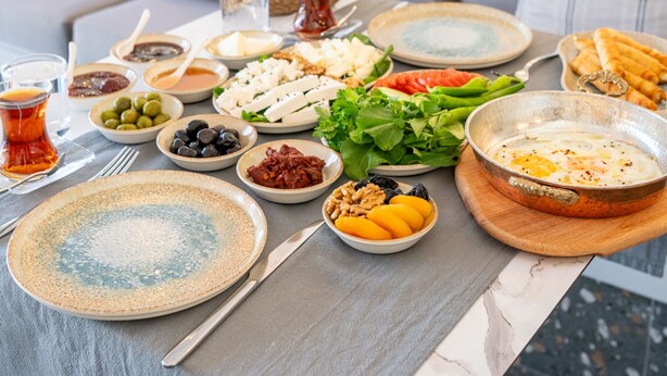 Une table à manger dressée avec différents plats et boissons, placée dans un salon moderne avec un canapé gris et des appliques murales décoratives.