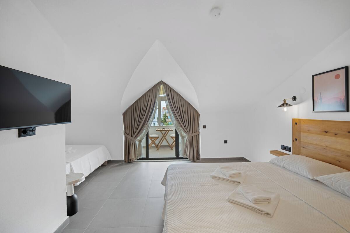 Una habitación de hotel moderna con una cama grande, televisión, una mesita y vistas a través de una ventana con cortinas.