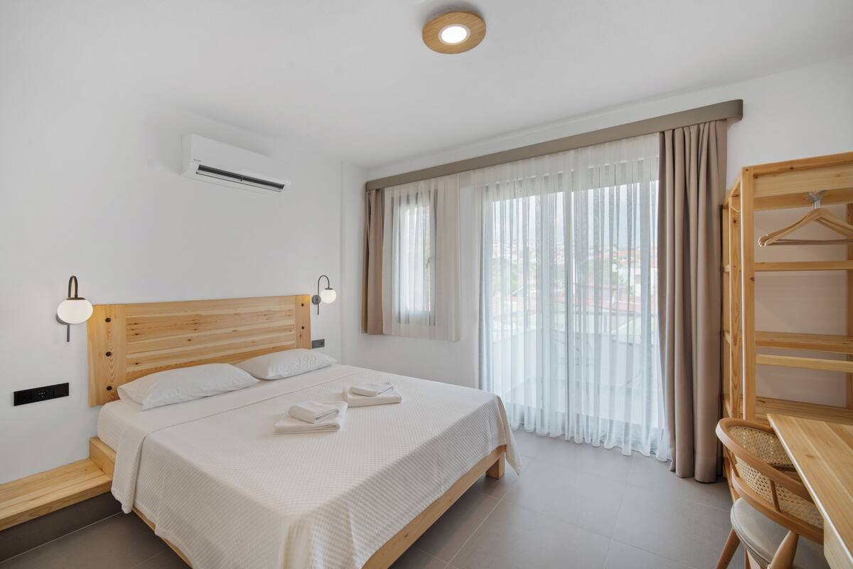 Un dormitorio con una cama de madera, una mesita de noche, una lámpara colgante y una ventana con cortinas translúcidas.