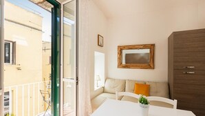 A balcony with a glass door, a white table with a potted plant, a beige sofa with an orange cushion, and a wooden cabinet.