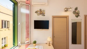 A room with a desk, chair, open window, air conditioning unit, flat-screen TV, decorative wall art, and a door.
