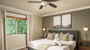 Un dormitorio con cama, ventilador de techo, ventana con cortinas, mesitas de noche y decoración en la pared.
