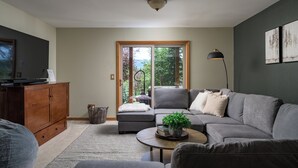 Una sala de estar moderna con un sofá gris, un mueble de madera, una mesa de centro con una planta y un ventanal con vistas a árboles.
