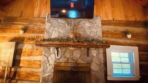 L’intérieur d’une cabane en rondins, avec une cheminée en pierre, un téléviseur fixé au mur et une fenêtre.