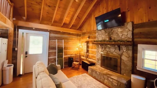 L’intérieur chaleureux d’un chalet en bois présente une cheminée en pierre, un téléviseur à écran plat fixé au-dessus, et un coin salon confortable avec un canapé.