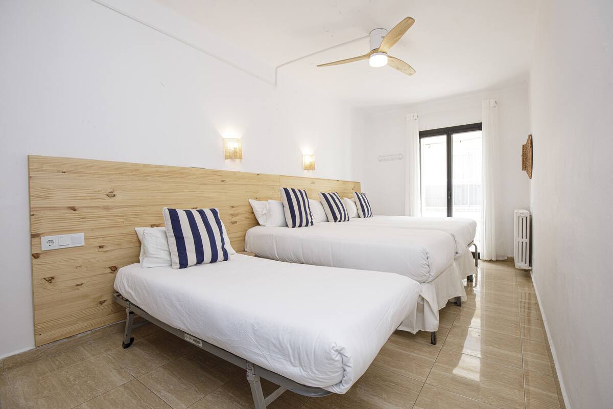 Ein Hotelzimmer mit zwei Betten, Holzkopfteilen, weißer Bettwäsche, Wandlampen, einem Deckenventilator und einem großen Fenster mit Vorhängen.