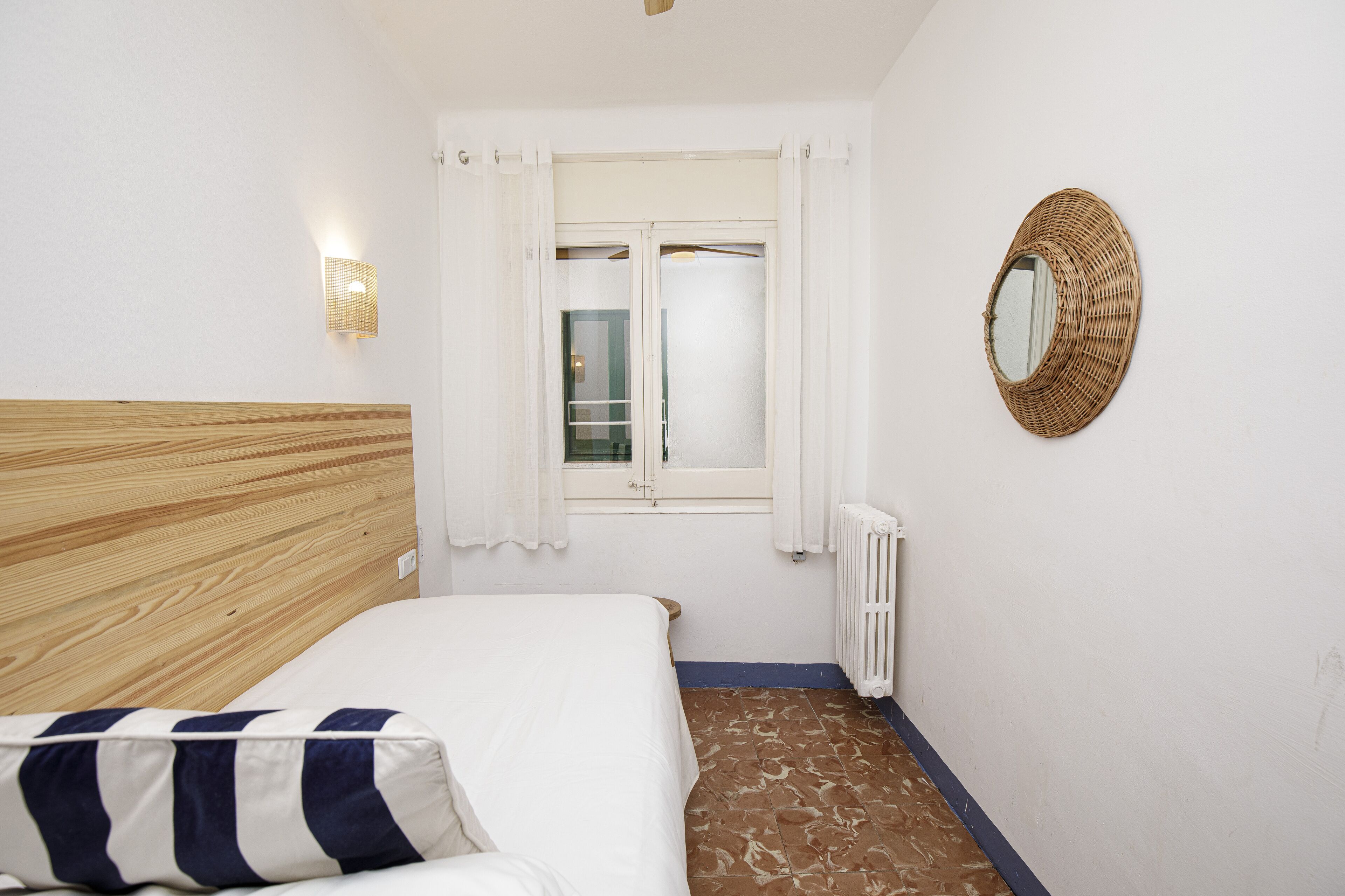 Un dormitorio con cabecera de madera, cama con almohada a rayas, ventana con cortinas blancas, espejo adosado a la pared y radiador.