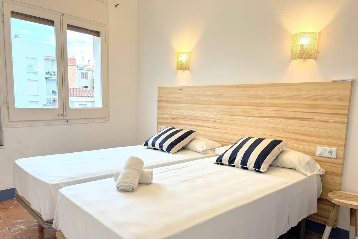 Ein Hotelzimmer mit zwei Betten, einem Holzkopfschild, gestreiften Kissen, einem kleinen Holztisch und einem Fenster mit Blick auf Gebäude.