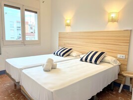 A hotel room with two beds, a wooden headboard, striped pillows, a small wooden table, and a window with a view of buildings.