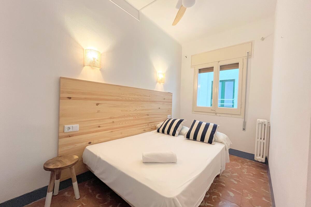 Ein Schlafzimmer mit einem hölzernen Kopfteil, zwei Nachttischlampen, einem Fenster, einer Heizung und einem kleinen Hocker.