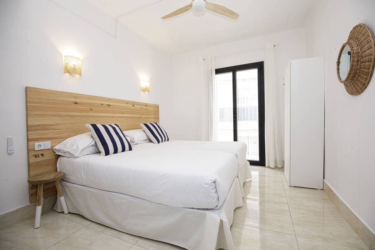 Ein Schlafzimmer mit einem Holz-Kopfteil, einem Bett mit weißer Bettwäsche, einem kleinen Holzschemel, einem weißen Kleiderschrank und einem Deckenventilator.