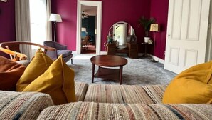 A living room with a purple wall, a sofa with yellow cushions, a wooden coffee table, and a mirror. - 1878 Victorian Charmer (Endicott)