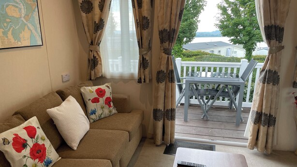A living room with a sofa, a table, and a view of a balcony with a chair. - Poppy, dog-friendly caravan with panoramic sea views on Rockley Park Poole wi-fi (Poole)