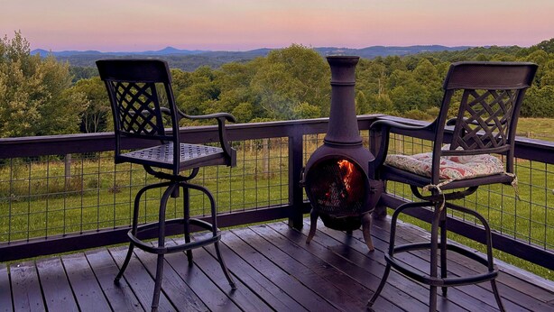 Terraza de madera con dos sillas de metal y una mesa, con vistas a un paisaje verde y un cielo despejado.