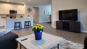A modern living room with a TV, a coffee table with a flower vase, a couch, and a kitchen area with a microwave and stools. - Country Cozy! Well stocked, immaculate home. Great short or extended stays!   
 (Jarrell)