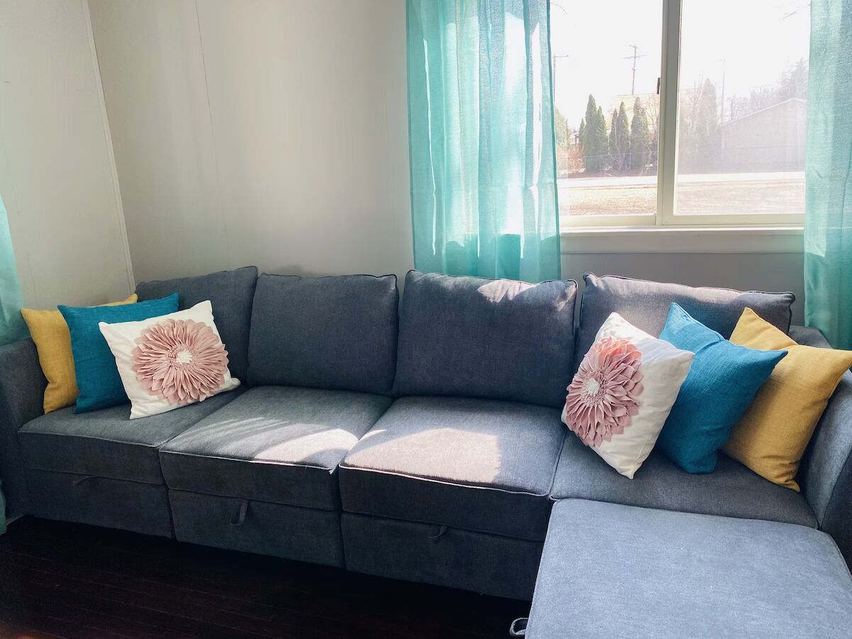 A grey sectional sofa with multiple cushions in various colors and a large window with sheer curtains.