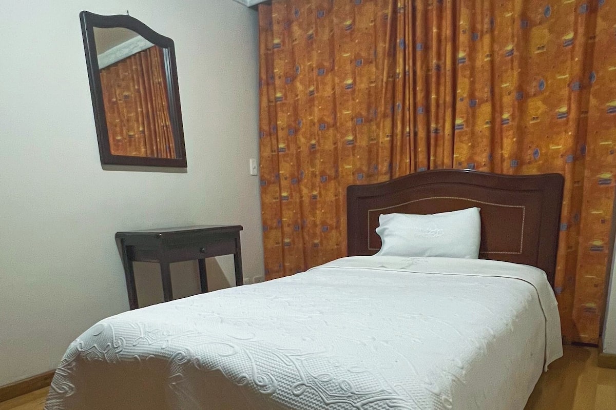 A bedroom with a wooden floor, a bed with a white bedspread, a wooden headboard, a nightstand, a mirror, and a patterned curtain.
