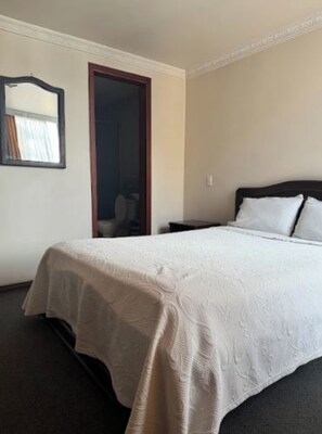 Habitación de hotel con una cama grande, una mesita de noche, un espejo y una puerta que da a un baño.