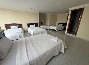 Habitación de hotel con cuatro camas individuales, cabecera de madera y baño visible a través de una puerta abierta.