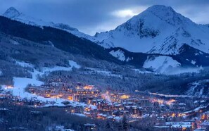 A snow-covered mountain town at dusk with illuminated buildings and a prominent mountain peak in the background.