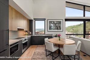 A modern kitchen with wooden cabinets, a round dining table with chairs, and a large window offering a view of the outdoors. - Charlie's Slopeside: Penthouse Ski-In/Out, Sleeps 10, Pool + Waterslide, Snowmass Base Village (Snowmass Village)