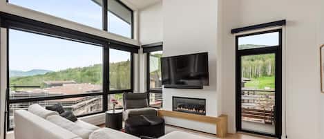 A modern living room with a sofa, a fireplace, a television, and large windows offering a view of the outdoors.