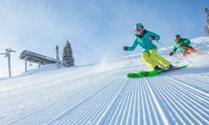 A ski slope with two skiers in motion, a ski lift in the background, and a clear sky.
