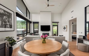 A modern living room with a round wooden table, grey sofas, a flat-screen TV, and a large window offering a view of the outdoors. - Charlie Rentz: Penthouse Ski-In/Out, Sleeps 10, Pool + Waterslide, Snowmass Base Village (Snowmass Village)