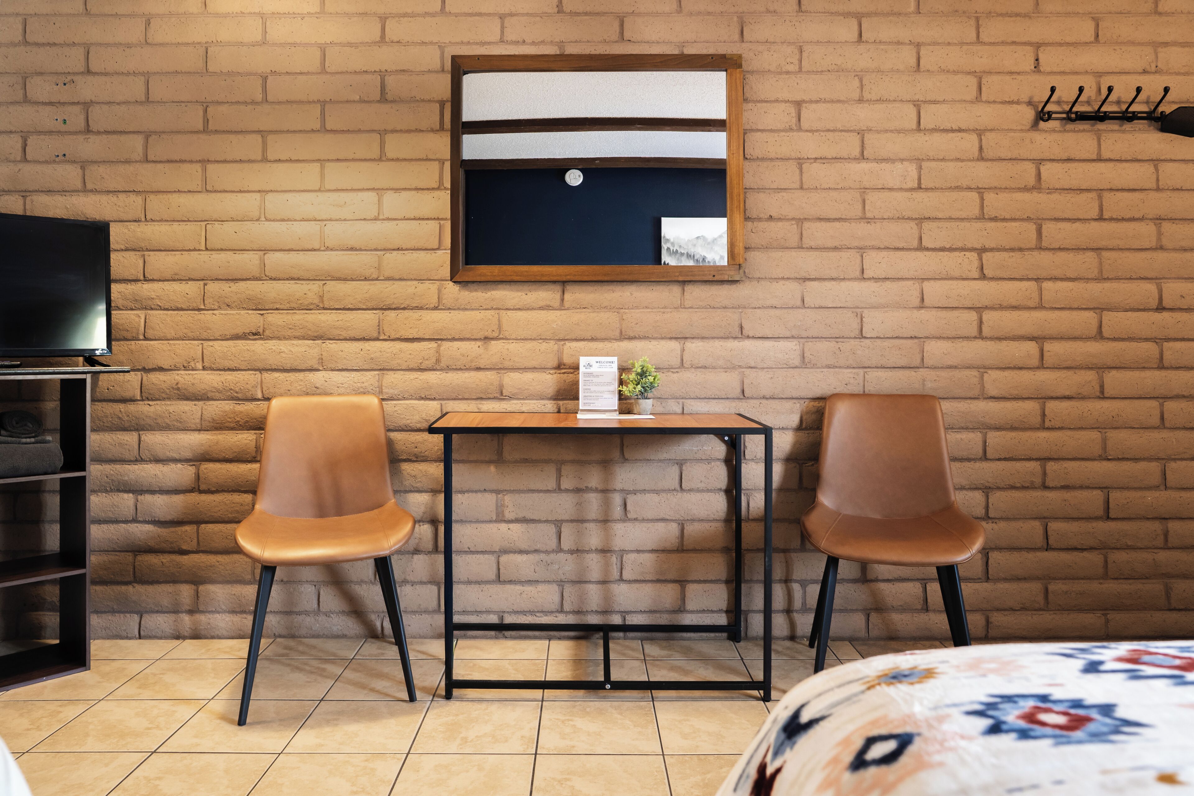 A room with a brick wall, a television, two chairs, a small table with a plant, and a bed with a patterned bedspread.