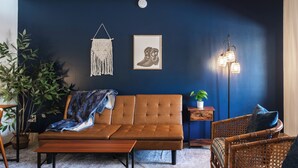 A room with a dark blue wall, a brown sofa, a wicker chair, a small side table with a plant, and a hanging macrame piece.