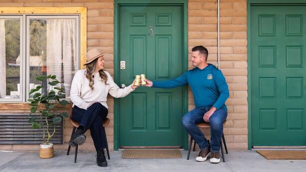 Two people sitting on chairs outside a building with green doors, one holding a bottle.