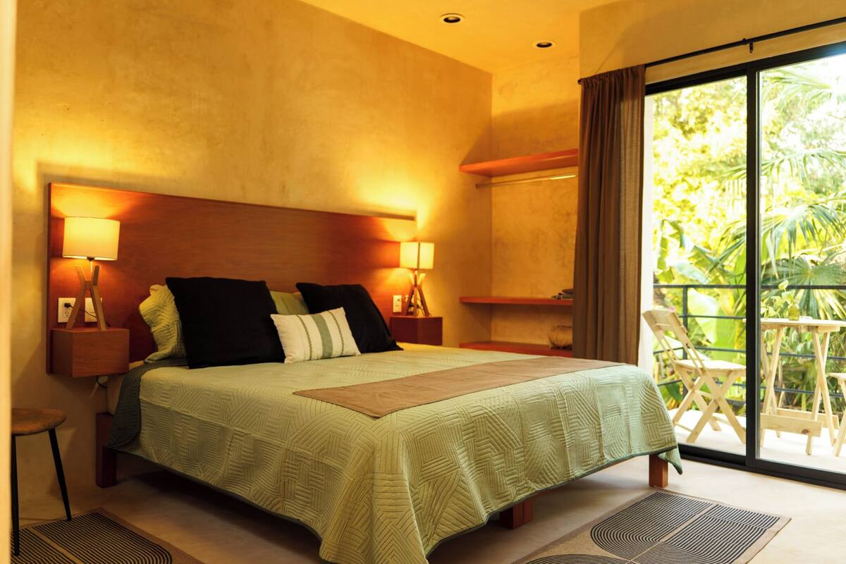 A hotel room with a large bed, two bedside lamps, a nightstand, a chair, and a view of greenery through a sliding door.