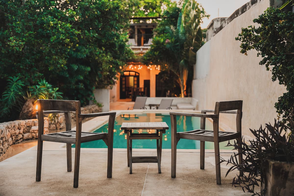 A poolside area with two wooden chairs and a small table, surrounded by lush greenery and a stone wall.