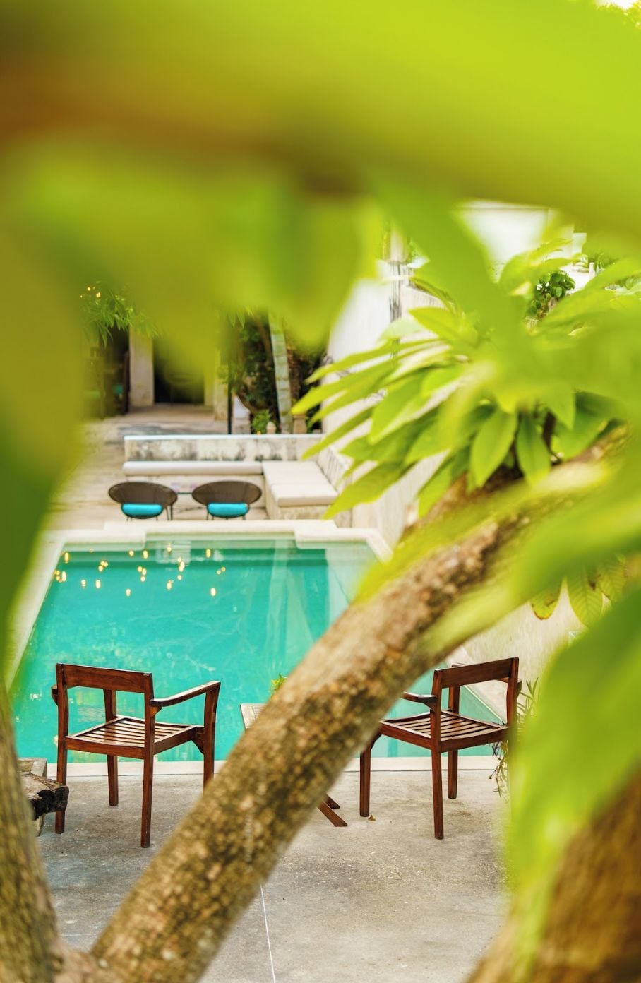 A pool area with two wooden chairs and a small table, surrounded by greenery.