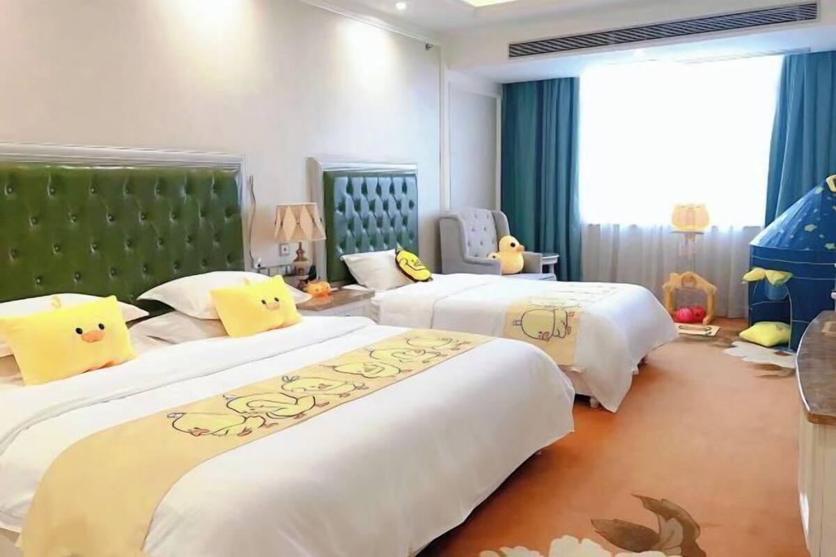 A hotel room with two beds, a green upholstered headboard, a chair, a lamp, and a window with curtains.