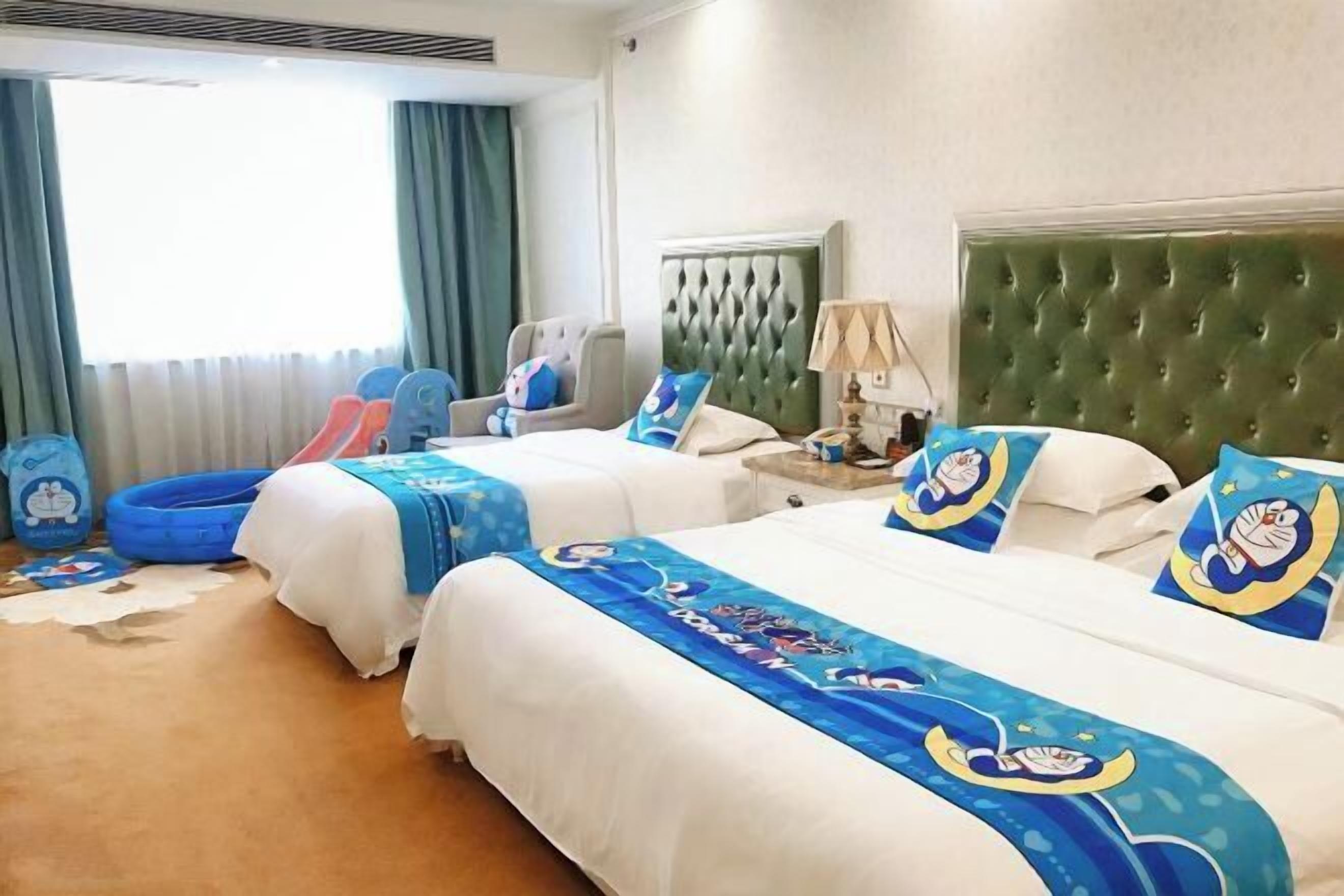 A hotel room with two beds, a green tufted headboard, a blue and white themed bedspread, a nightstand with a lamp, and a window with curtains.