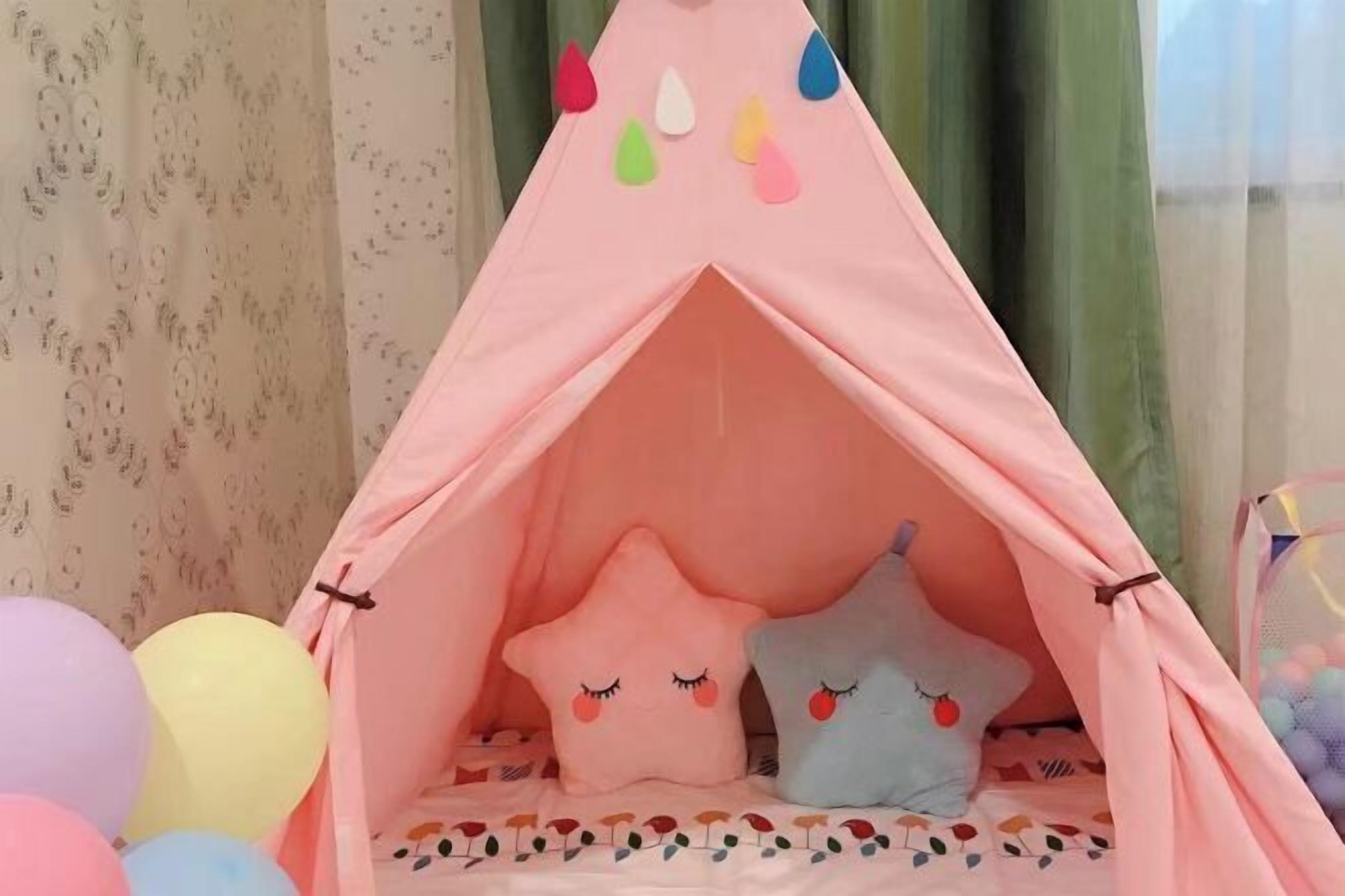 A pink tent-like structure with colorful teardrop decorations, two plush pillows with cute faces, and a bed with a patterned bedspread.