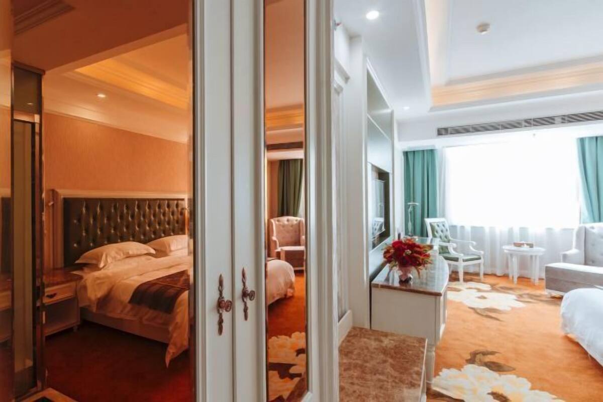 A hotel room with a bed, a sitting area with a chair and a table, a mirror, and a large window with curtains.