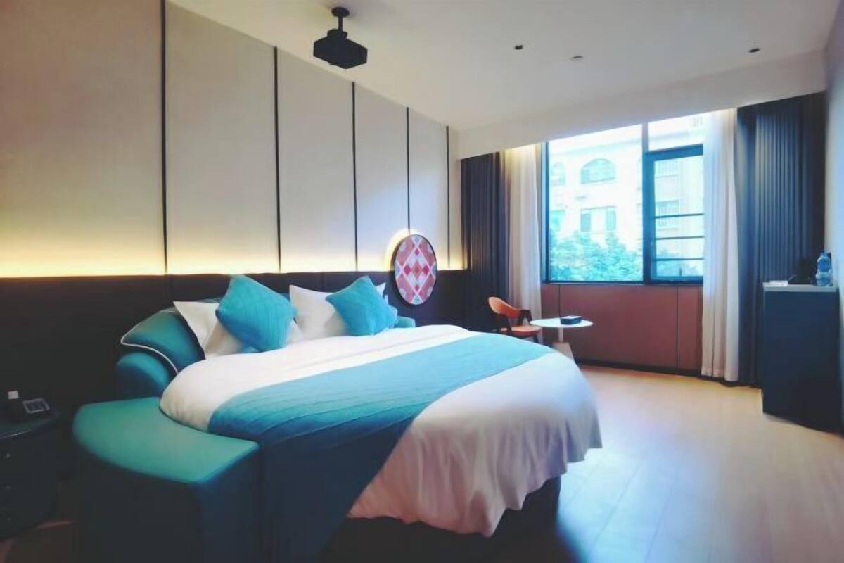 A modern hotel room with a large bed, a small table, a chair, and a view of a building outside.
