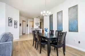 A dining area with a dark wooden table, six chairs, and a chandelier. There are framed artworks on the walls and a gray sofa in the foreground. - Epic Universe Penthouse Vacation Condo, Near the Parks - 2014 (Orlando)