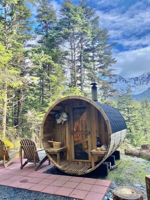 A wooden barrel-shaped cabin with a small porch and seating area, surrounded by a forest and mountains in the background. - Little Bear Den - Nice Eco-Studio + Outdoor space (Seward)