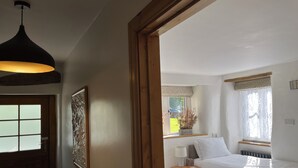 A bedroom with two beds, a bedside table with a lamp, a window with a view of the outside, and a ceiling light fixture. - The Yorkshire Dales Snug - Walkers' Delight - Pet Friendly (Buckden)