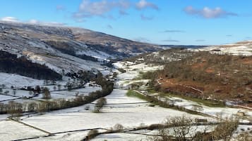 Paisagem rural com campos cobertos de neve, uma estrada sinuosa e colinas ao longe.