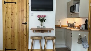 A compact kitchen with a wooden door, a mounted TV, a small wooden shelf with a vase of flowers, and a microwave above the counter. - The Yorkshire Dales Snug - Walkers' Delight - Pet Friendly (Buckden)