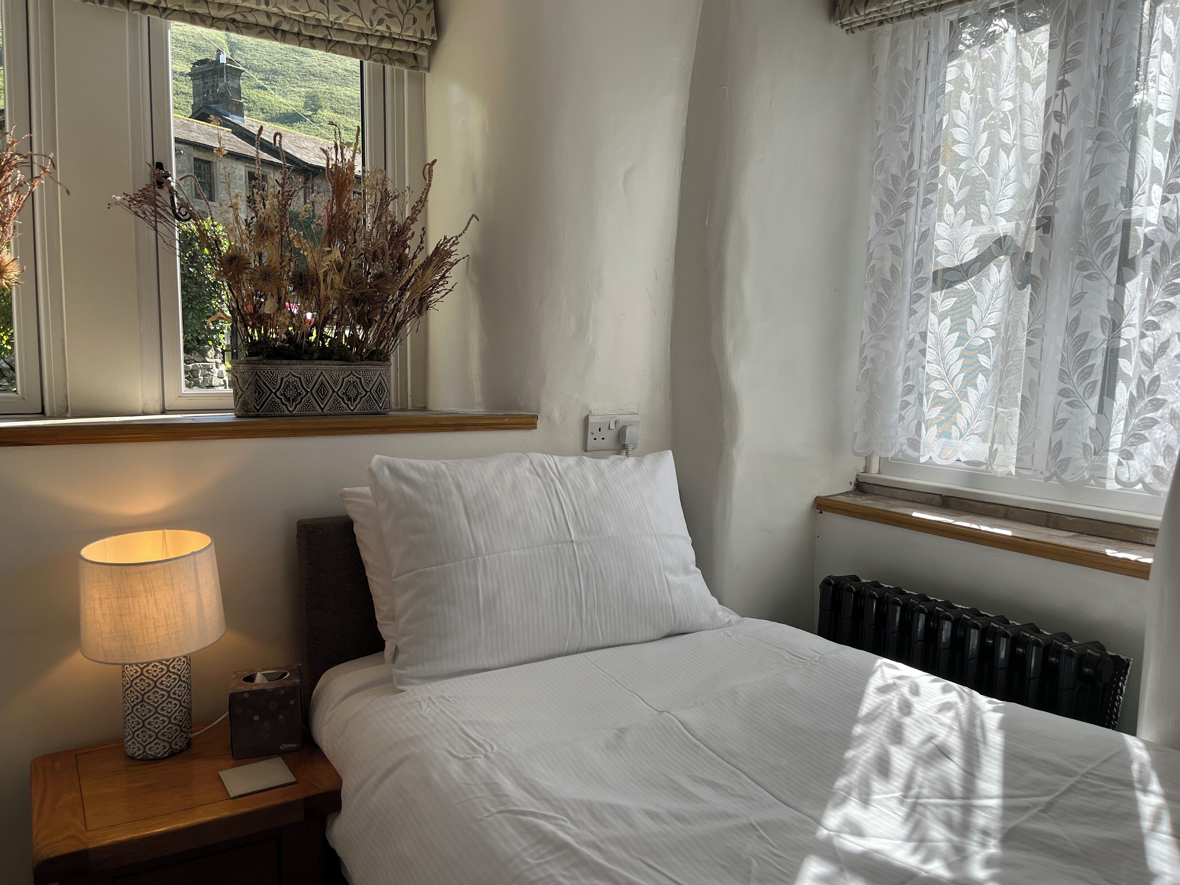A bedroom with a bed, a nightstand with a lamp, a potted plant, and a window with curtains.